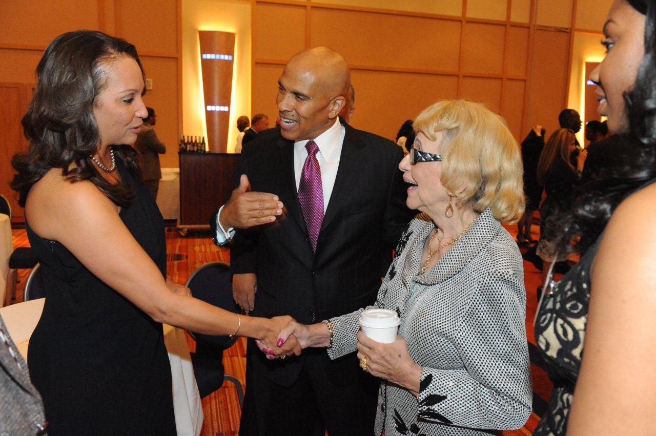 Four people converse at a formal event. Two women shake hands while a man in a suit speaks with them, and another woman stands nearby.