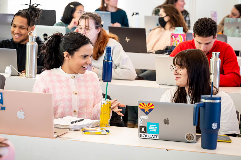 Law students work on laptops and talk with classmates in a classroom.