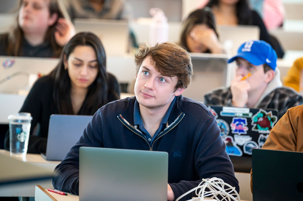 A law student listens attentively during a classroom lecture, with other students working on laptops in the background.