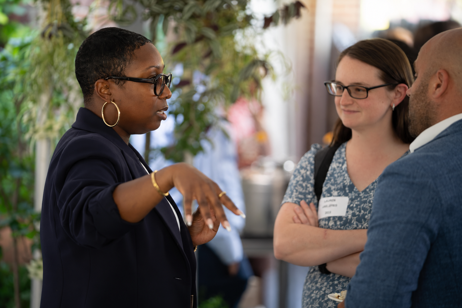Three alumni converse at an indoor networking event surrounded by greenery. A woman in the foreground gestures while speaking to two attendees.