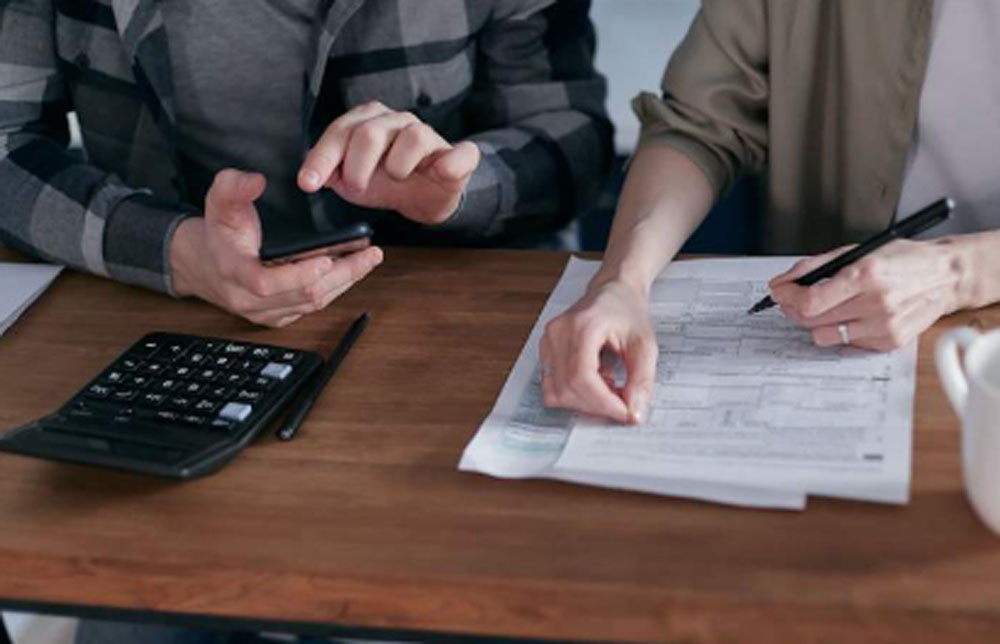 Two people review tax documents together while using a calculator and smartphone at a desk.