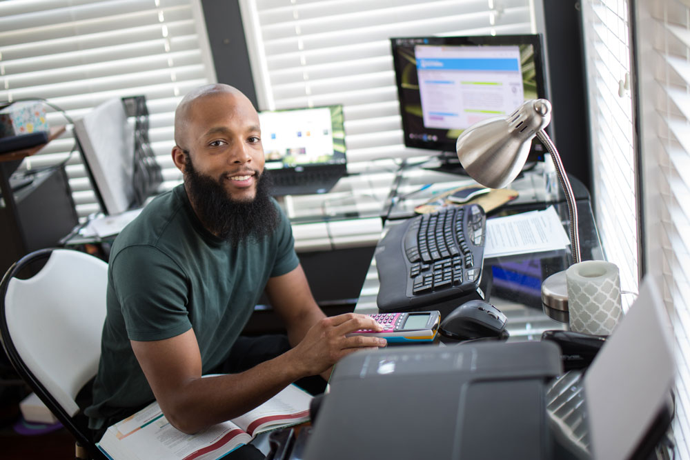 A student studies from a home office while using a calculator, computer, and course materials for online learning.