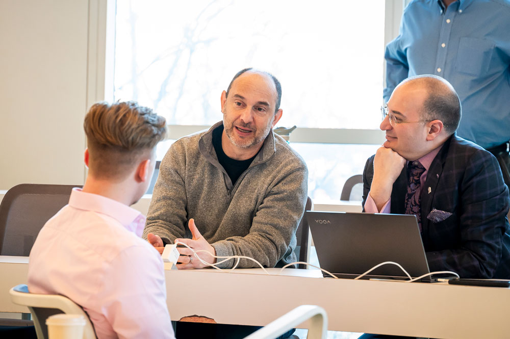 Three people seated around a table in conversation, one gesturing while speaking and another listening with a laptop open in front of him
