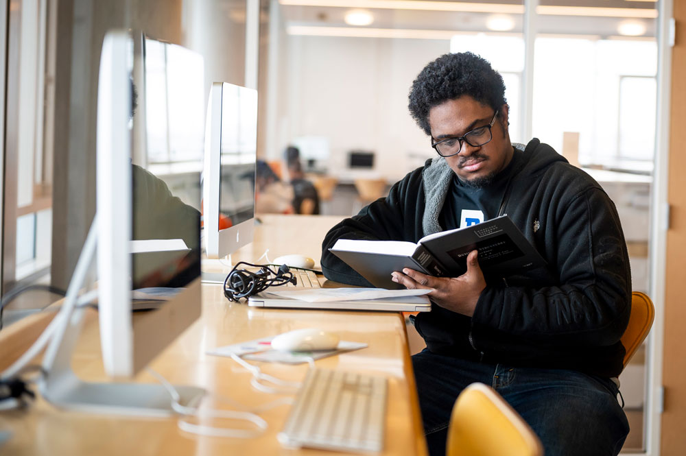 A student reading a casebook while seated at a computer workstation in a study area, with a laptop and papers open on the desk in front of him
