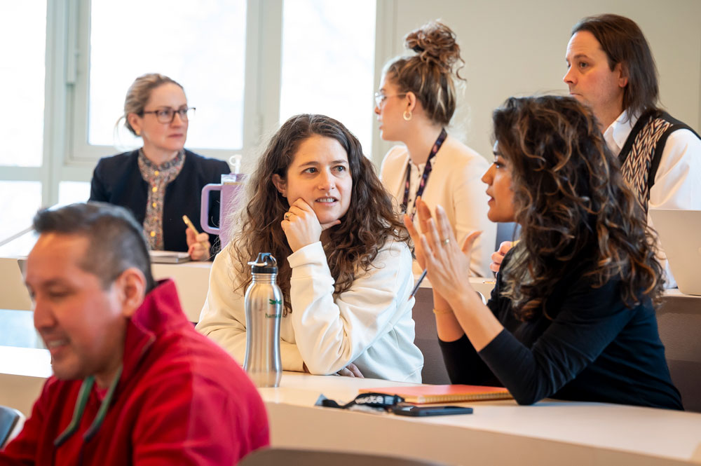 Faculty seated at tiered classroom desks during a discussion, one gesturing as she speaks while others listen and turn toward her