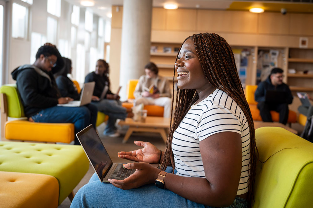 A student smiling and talking with a laptop open on her lap in a bright lounge, with other students working on couches behind her