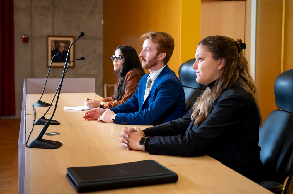 Three students in professional attire seated at a long table with microphones in a moot courtroom, looking ahead attentively