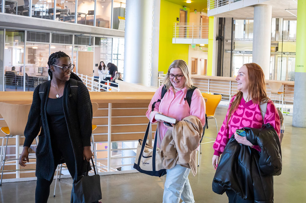 Three students walking and talking together in the Angelos Law Center atrium, carrying bags, coats, and books