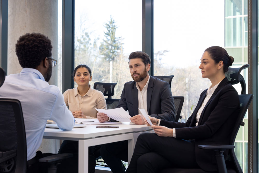 Four professionals meet around a conference table in a bright, modern office, with several participants holding documents.
