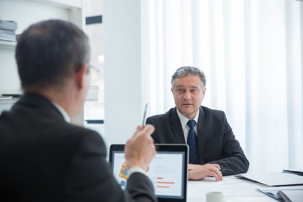 Two professionals meet across a desk to discuss documents and business matters in an office.