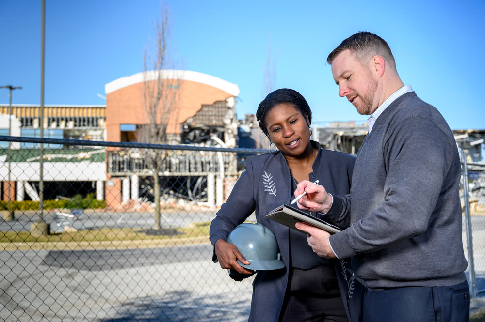 Two professionals review project plans on a tablet while standing at an active construction site.