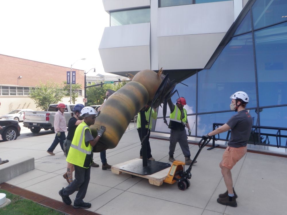 A large, realistic sculpture of a honeybee is being carefully moved outdoors. The honeybee sculpture is mounted on a sturdy black pedestal that sits upon a pallet. On person is pulling it using a pallet jack while other people are present, assisting with the relocation process; some are guiding the sculpture while others observe. 