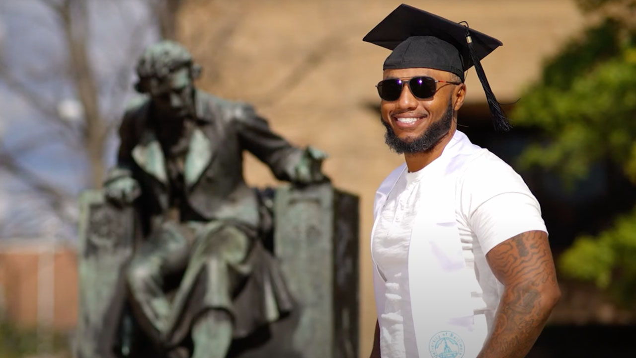A graduate in a graduate's cap and stole poses, smiling, in front of a statue of Edgar Allan Poe.
