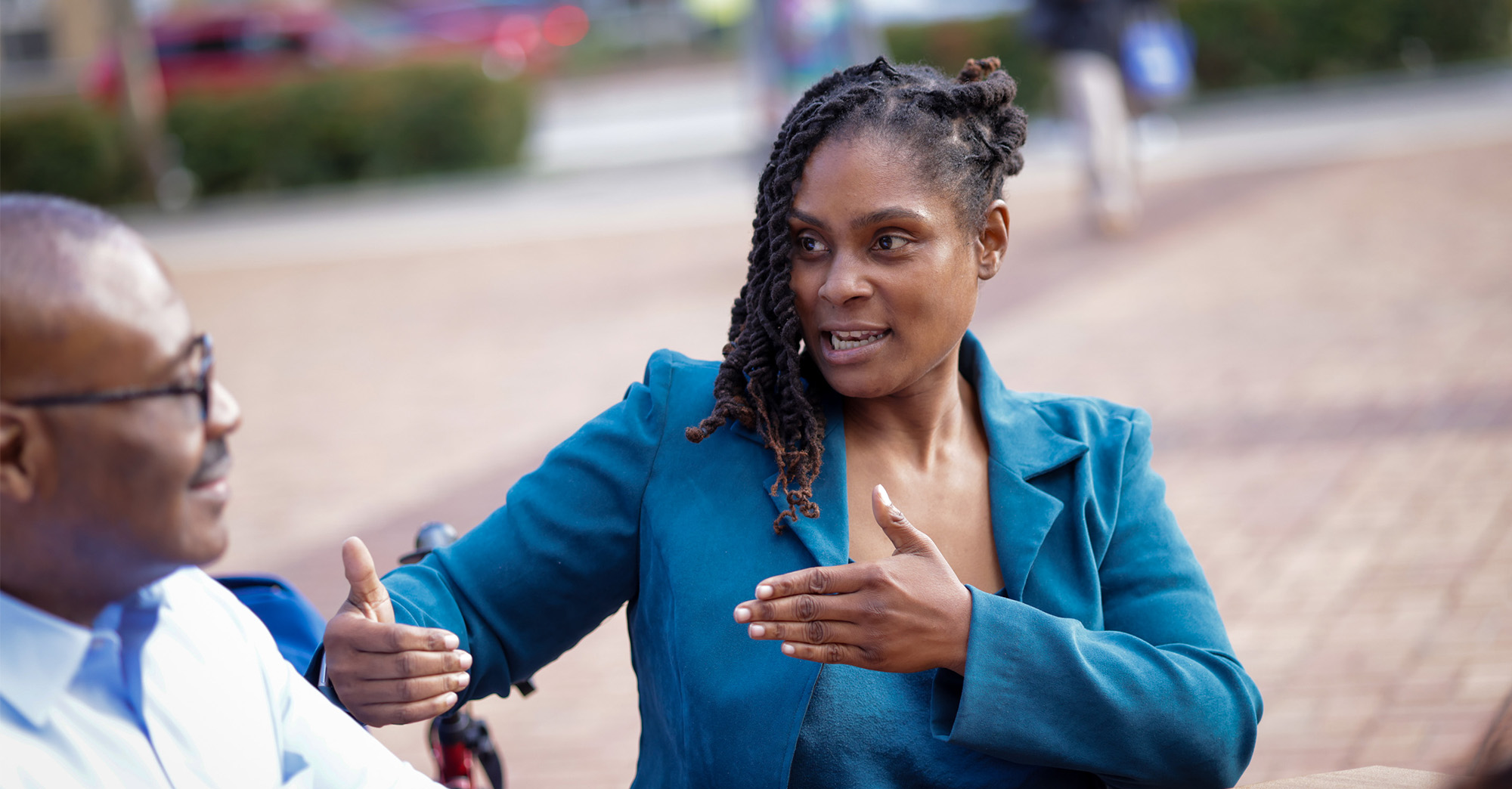 The image shows two people having a conversation outdoors, likely in an urban setting with a brick walkway and blurred background elements like greenery and passersby.