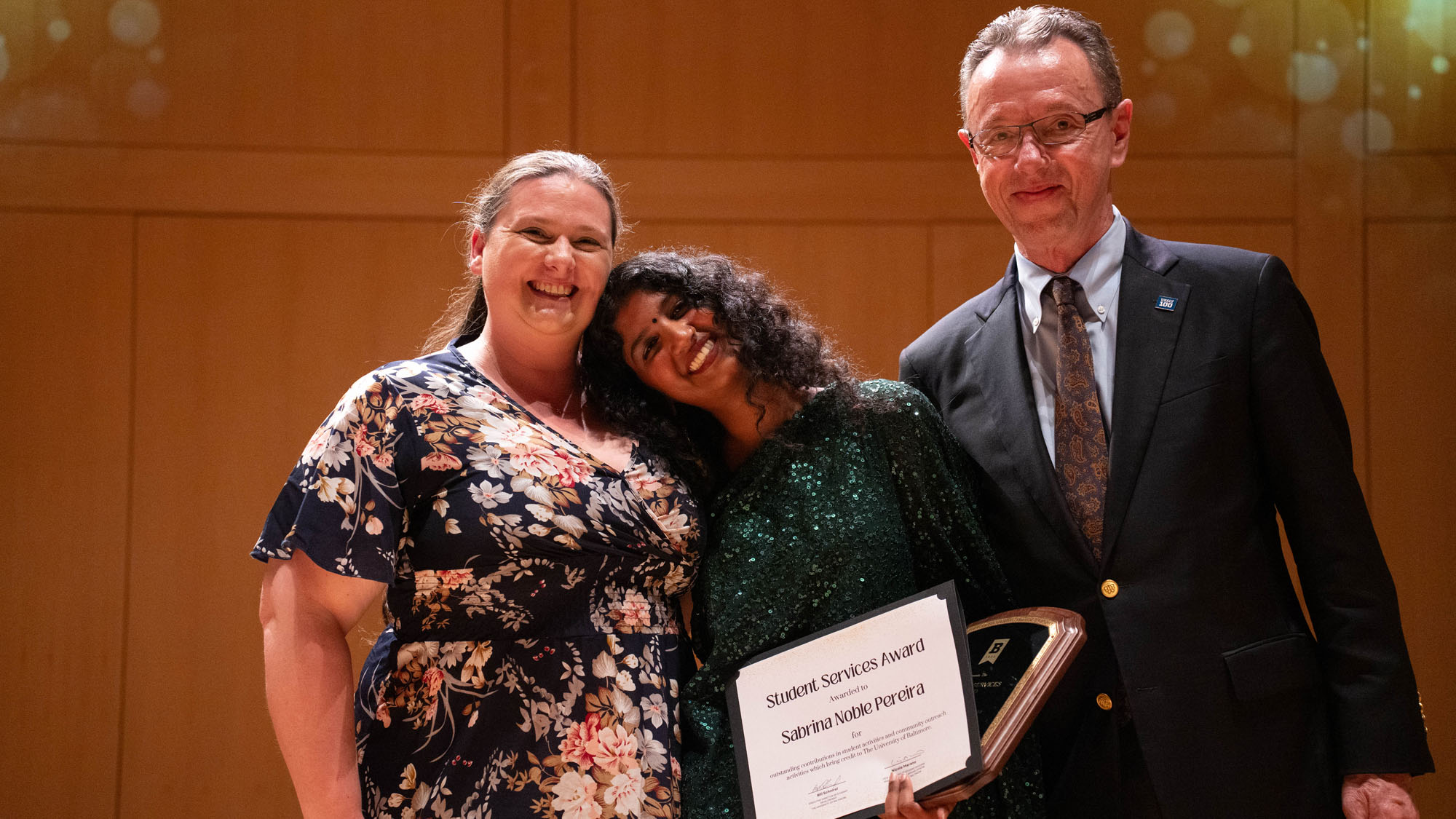 a student leans their head on another student's shoulder while both pose with an award presenter, all smile at the camera