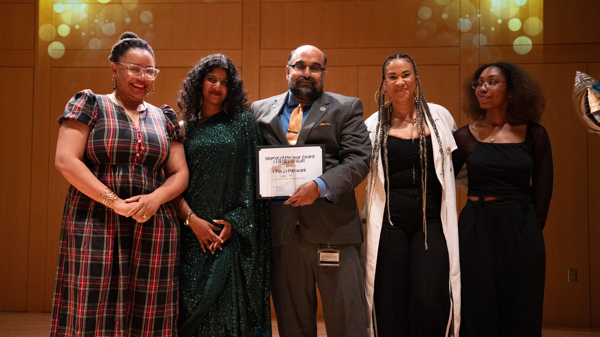 Five people stand on stage posing after the one at center holds the award they one; one looks fondly at the winner